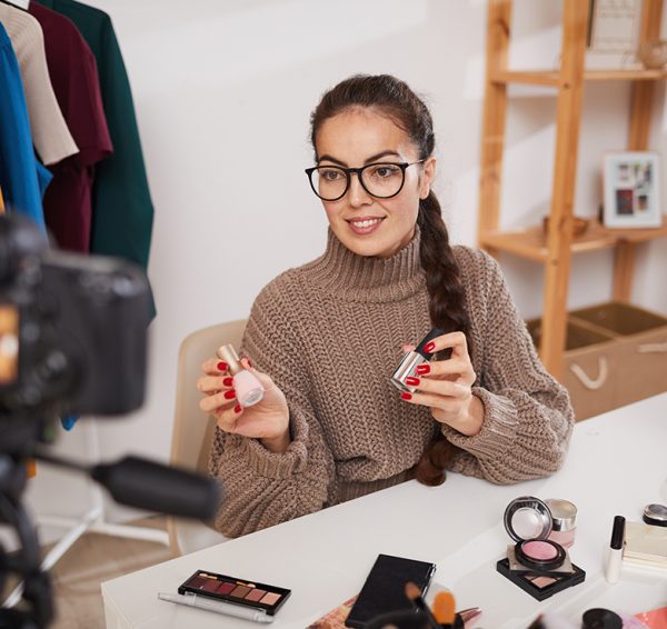 Portrait of smiling young woman showing makeup products to camera while filming review video for beauty and lifestyle channel, copy space