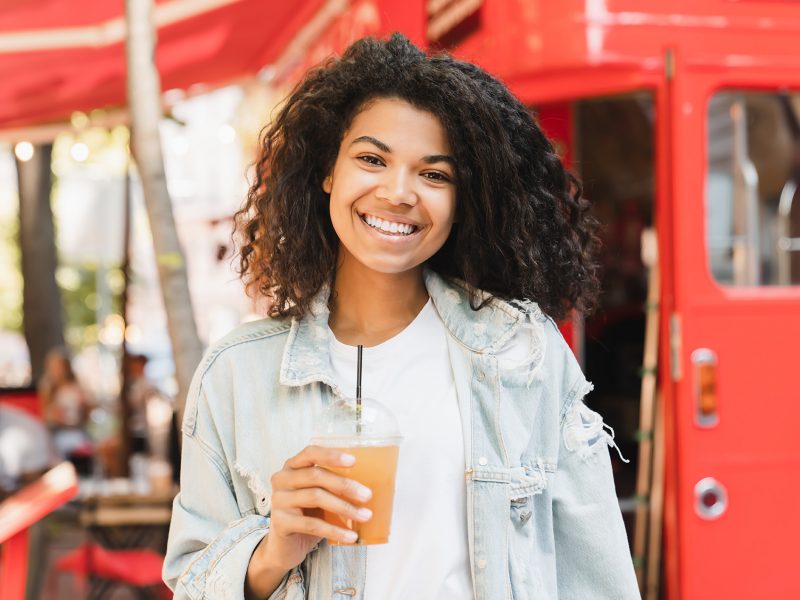 Young african-american woman businesswoman student drinking lemonade fresh juice walking in city park cafe outdoors. Portrait of an african female looking at the camera.